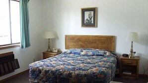 A bedroom with a large bed, two bedside tables with lamps, a wooden headboard, a framed picture on the wall, and a window with curtains. - The Skyline Motel (Osage Beach)