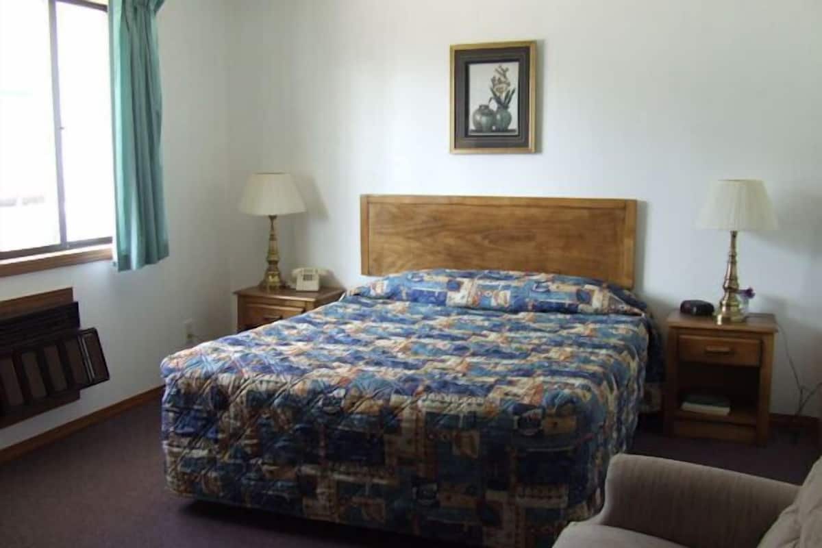 A bedroom with a large bed, two bedside tables with lamps, a wooden headboard, a framed picture on the wall, and a window with curtains.