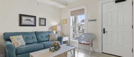 A living room with a blue sofa, a white coffee table, and a wicker chair. There is a white door and a window with a view of the outside.