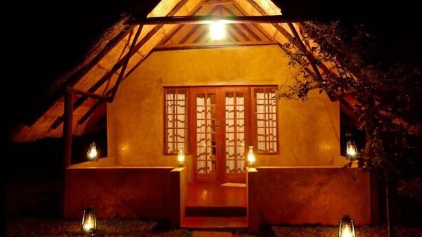 Estructura con techo de paja, puerta y ventanas de madera, iluminada por luces empotradas.