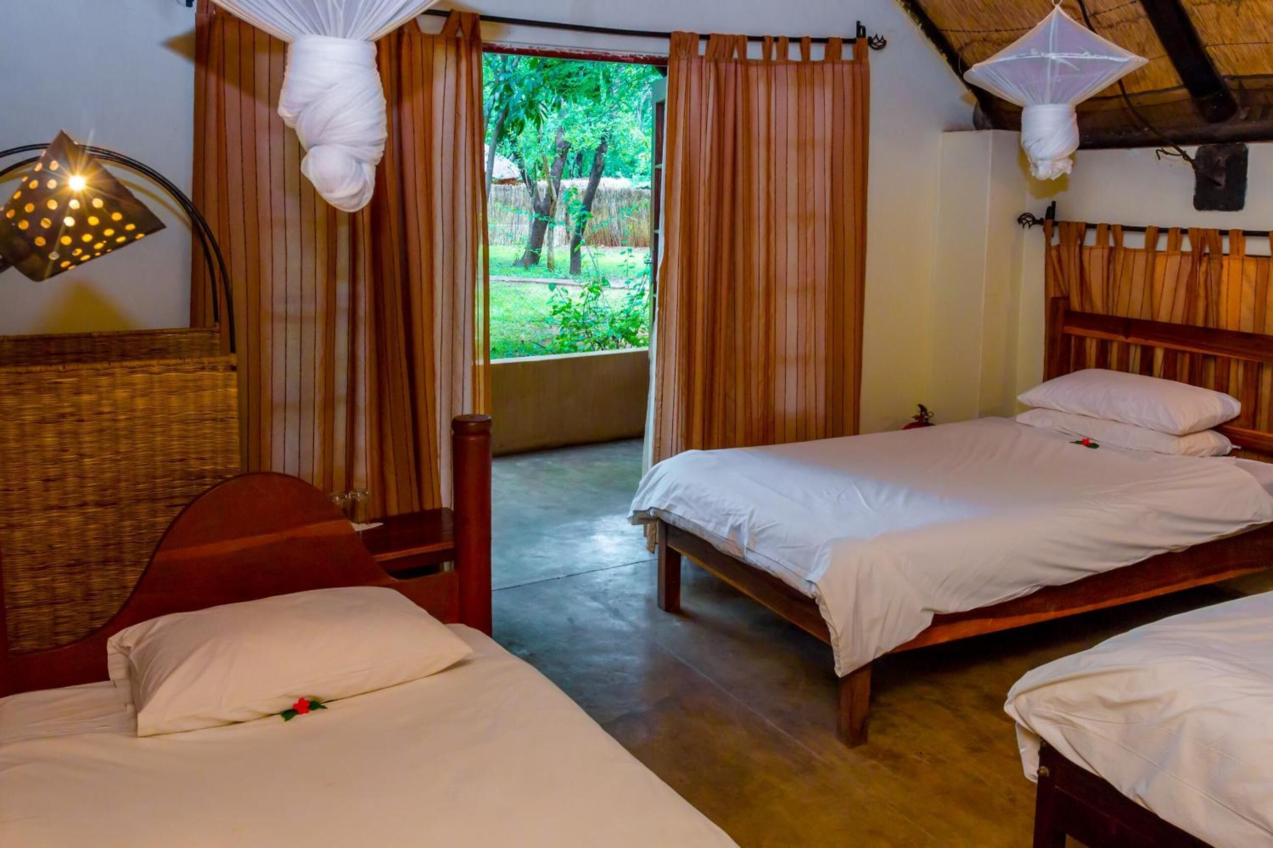 A room with two beds, a wooden headboard, a wicker chair, and a view of greenery outside.