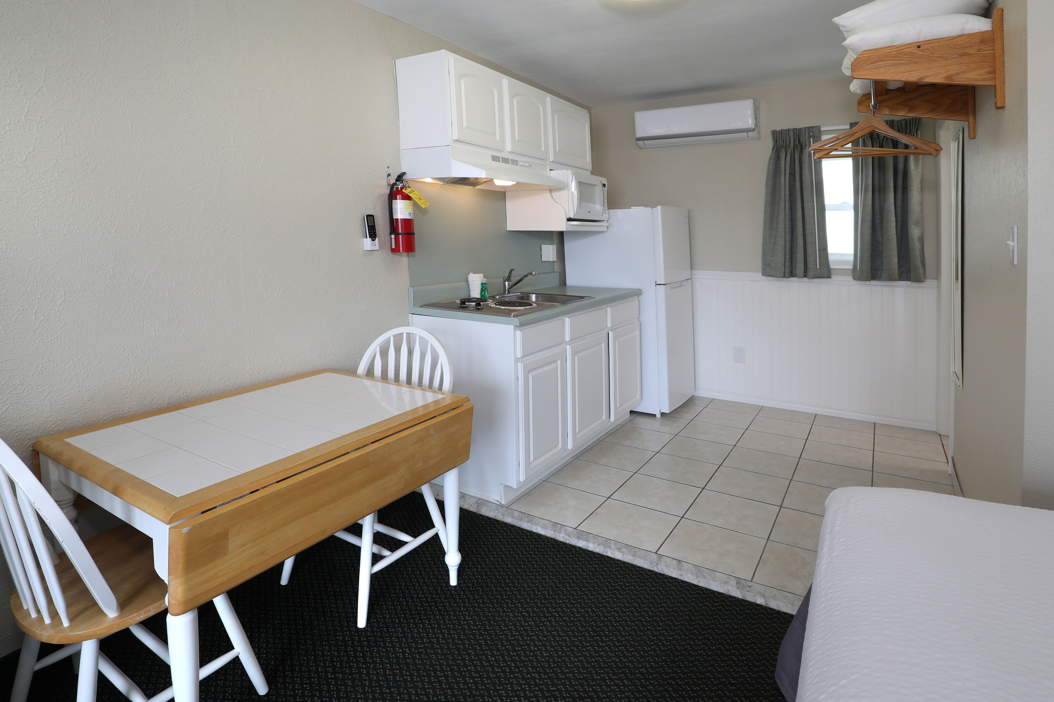 A compact kitchenette with a microwave, sink, and white cabinets. There is a small dining table with chairs and a bed in the room.