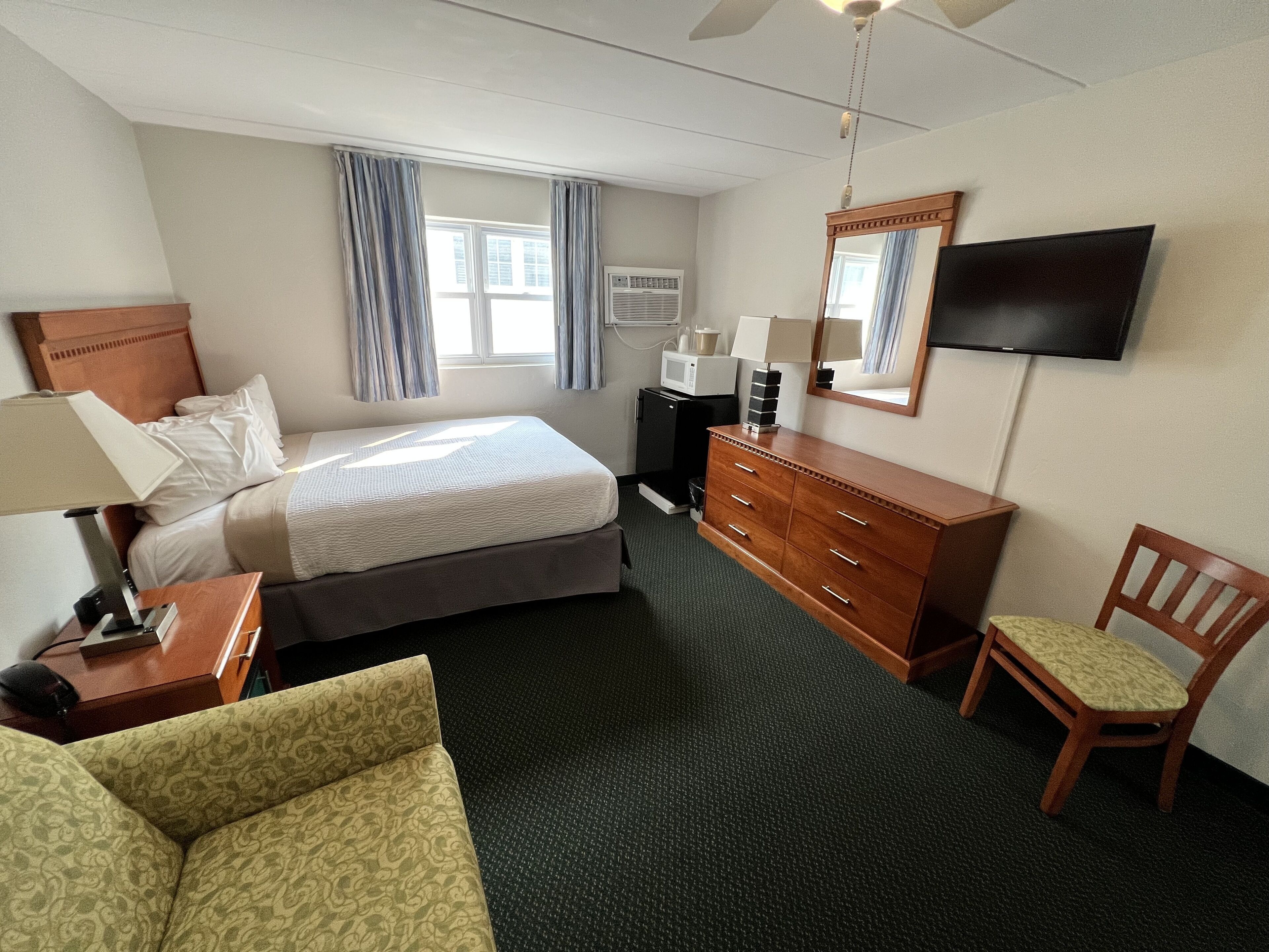 Habitación de hotel con cama, cómoda de madera, escritorio con lámpara, silla, televisor y ventana con cortinas.