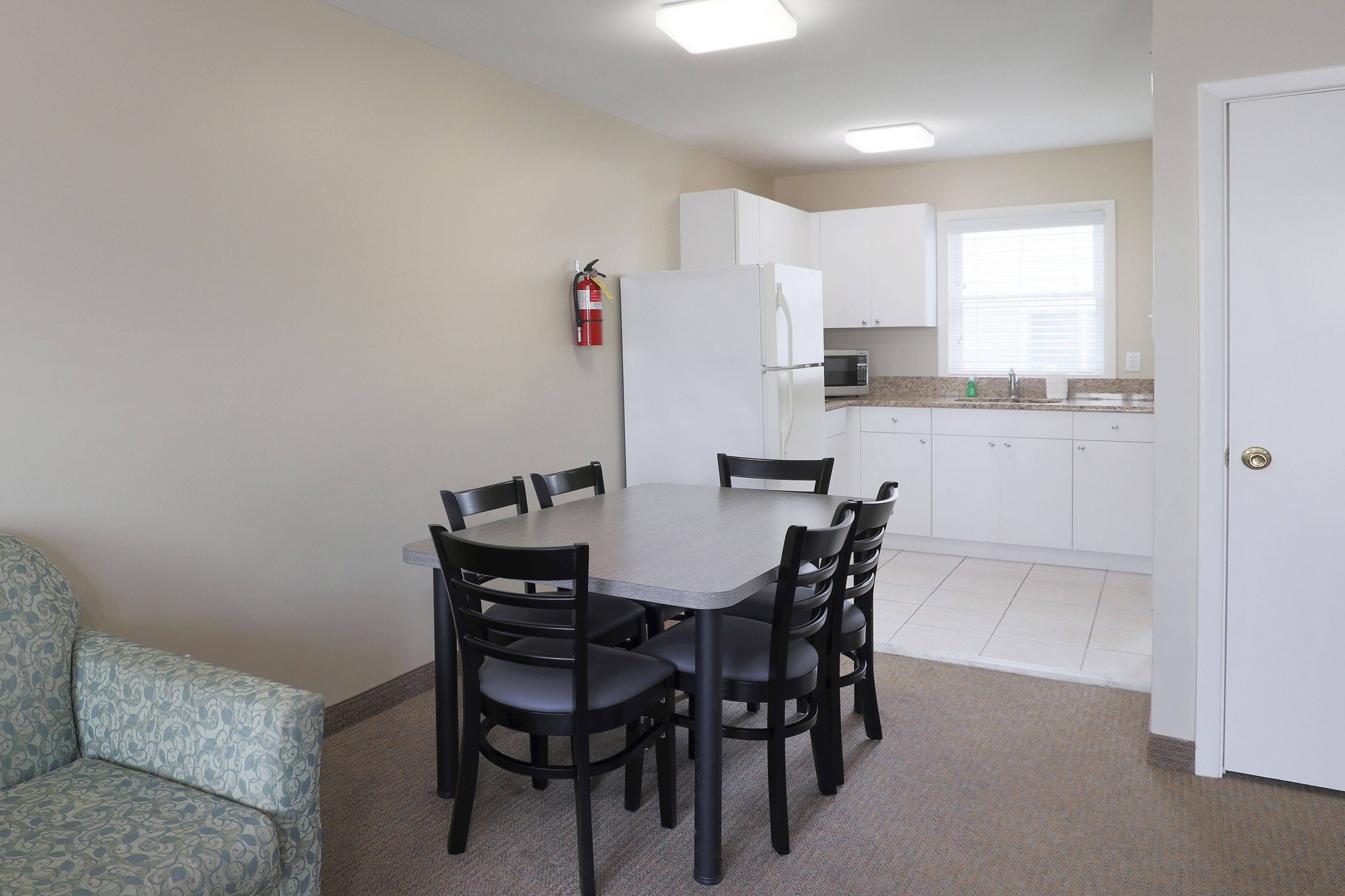 A compact kitchen with white cabinets, a refrigerator, and a microwave. There is a dining table with chairs and a fire extinguisher mounted on the wall.