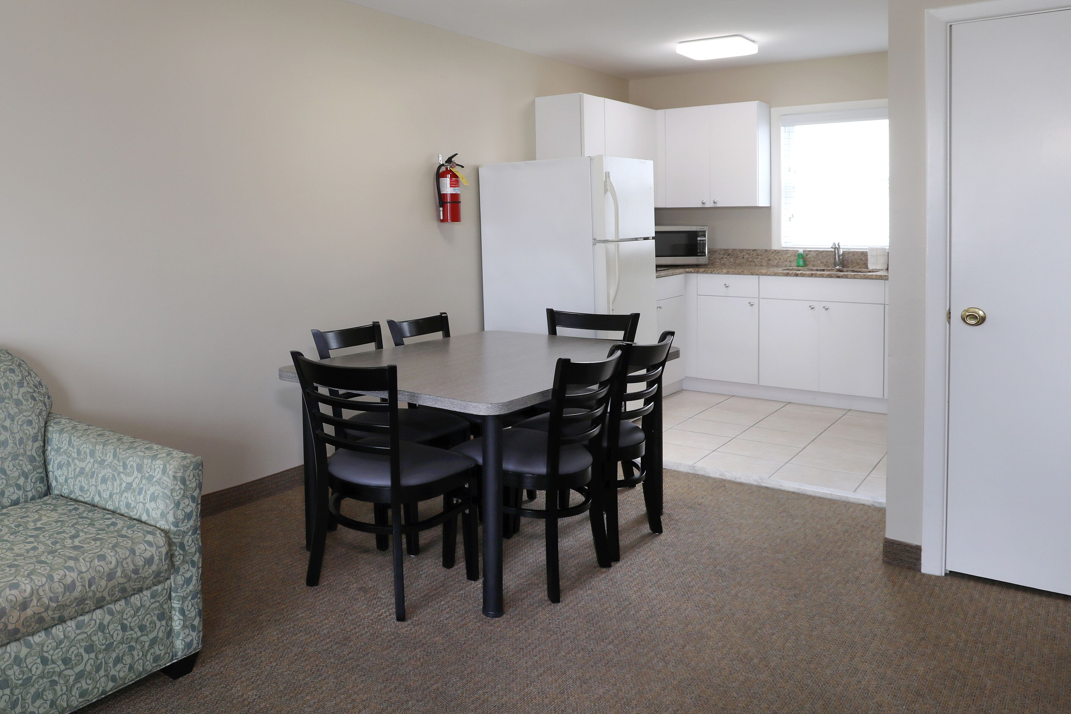 A compact kitchen with white cabinets, a microwave, and a refrigerator. There is a dining table with four chairs and a fire extinguisher mounted on the wall.