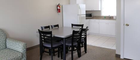 A compact kitchen with white cabinets, a microwave, and a refrigerator. There is a dining table with four chairs and a fire extinguisher mounted on the wall.