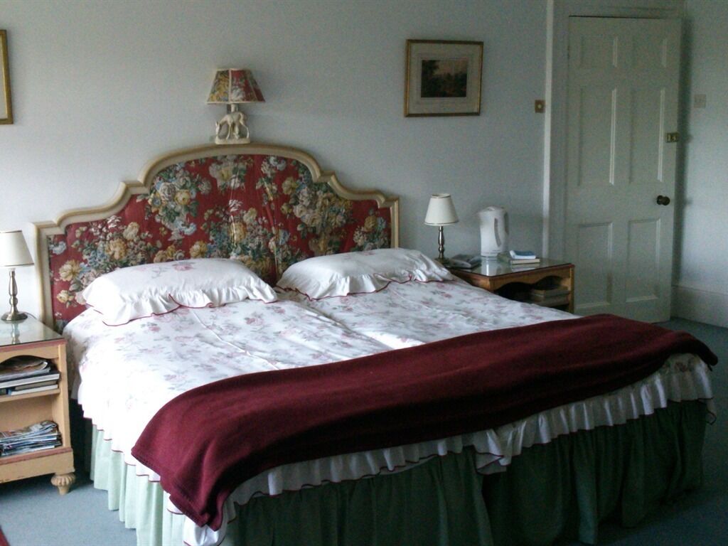 A neatly made bed with floral bedding and a red throw blanket. There is a nightstand with a lamp and a book beside the bed. A door is visible in the background.