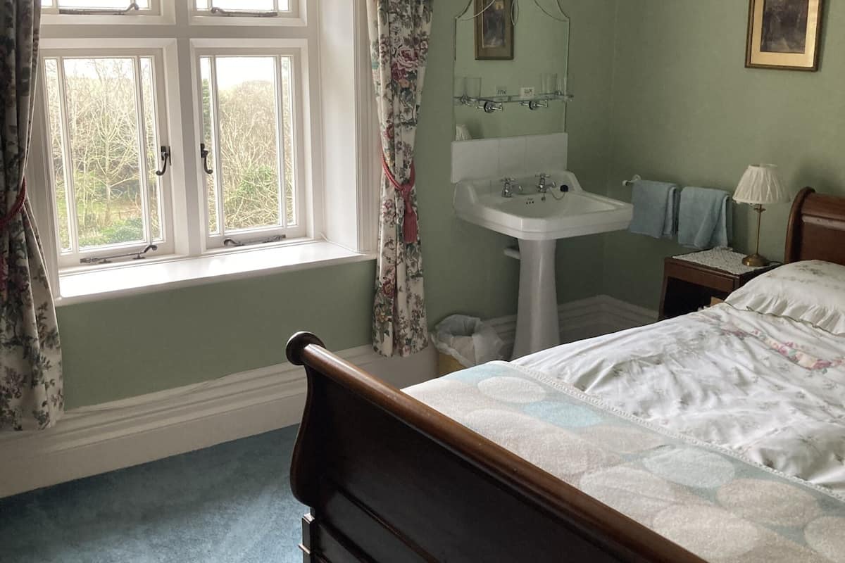 A bedroom with a bed, a window with curtains, a bedside table, a nightstand, a mirror, and a wall-mounted light fixture.