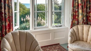 Une pièce dotée de trois fenêtres, d’un fauteuil beige, d’un tapis à motifs et offrant une vue sur la verdure et la mer.