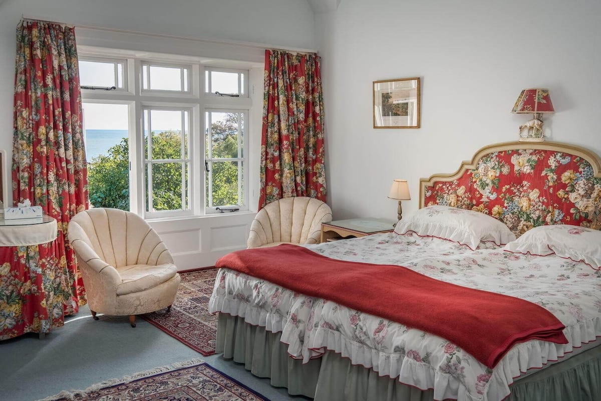 A bedroom with a bed, two chairs, a nightstand, a lamp, and a window with curtains.