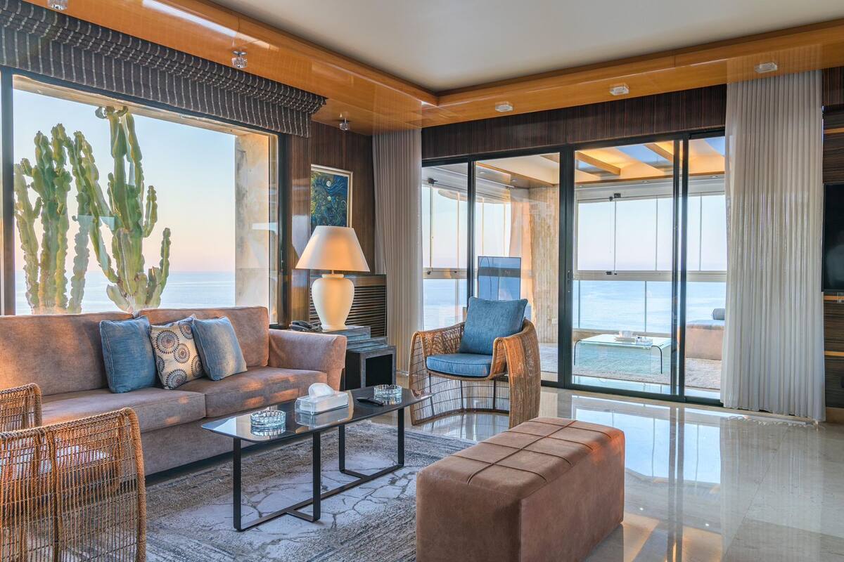 A modern living room with a sofa, armchair, and ottoman, a coffee table, and a lamp. There are large windows with a view of the ocean.