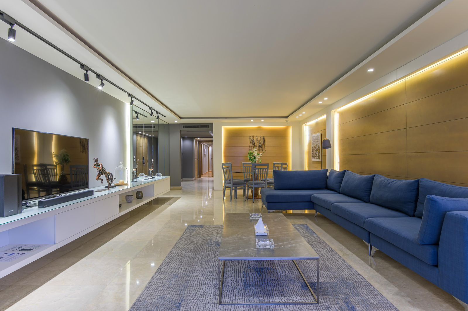 A modern living room with a large flat-screen TV, a blue sofa, and a glass coffee table.
