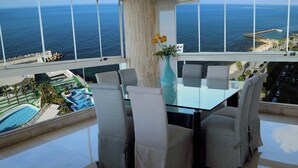 En matplats med ett glasbord och stolar, en vas med blommor och utsikt över havet och stranden.