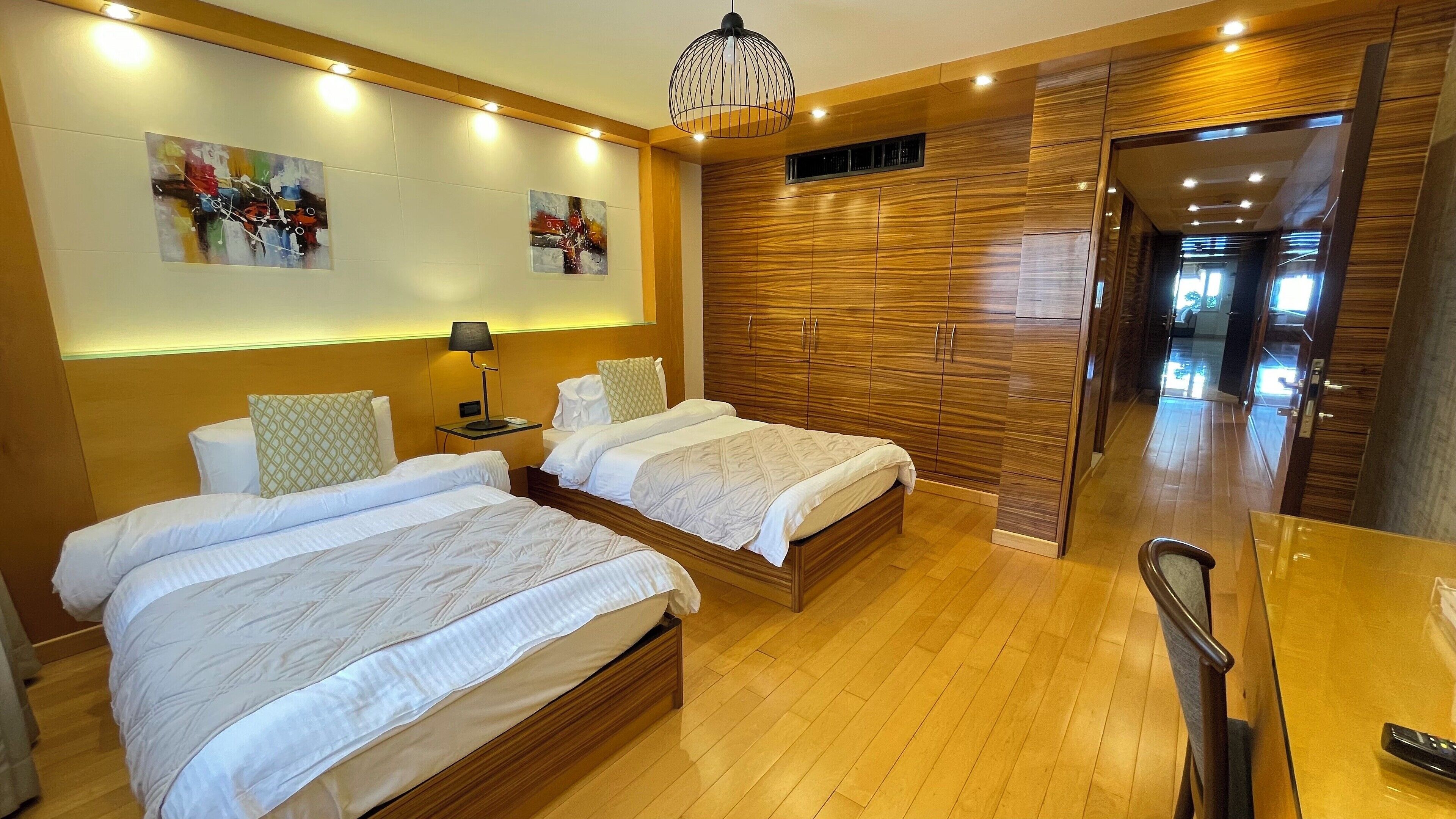 Une chambre d’hôtel avec deux lits, des têtes de lit en bois, une table de chevet avec une lampe, une armoire en bois et un couloir menant à une autre pièce.