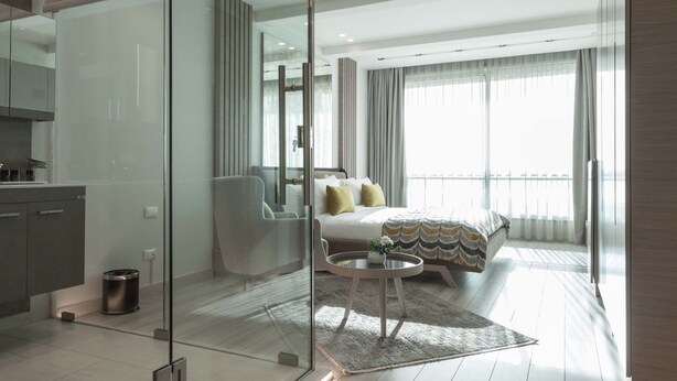 A modern hotel room with a glass partition separating the bedroom from the living area. The room features a large bed, a round coffee table, and a view of the city through large windows. - Lahoya Homes (Beirut)
