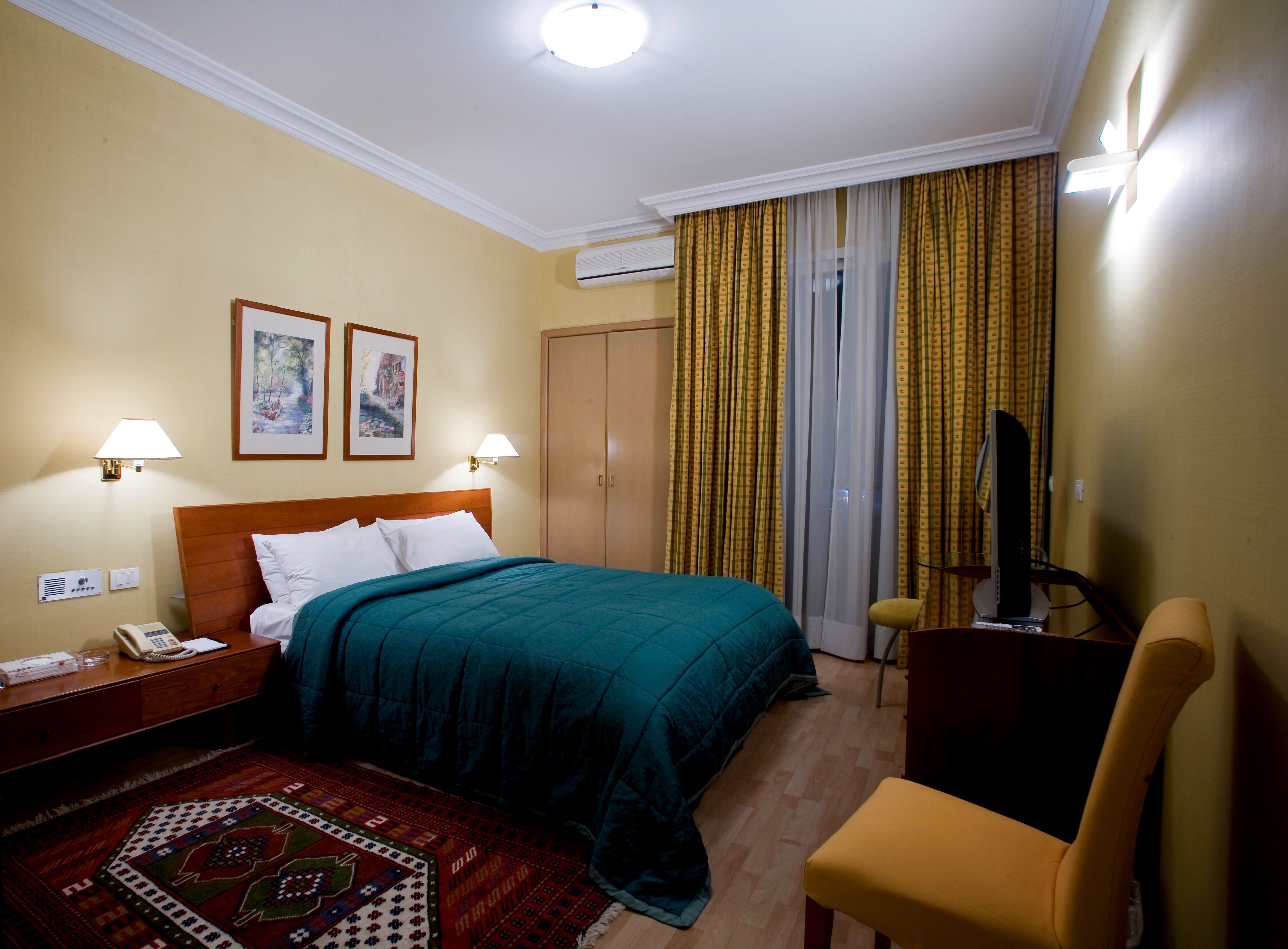 A hotel room with a bed, two wall-mounted lamps, a television, a desk with a telephone, and a patterned rug.