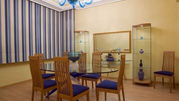 Un comedor con mesa de vidrio, sillas de madera con cojines azules, una araña, una cortina a rayas y un aparador de vidrio con objetos decorativos.