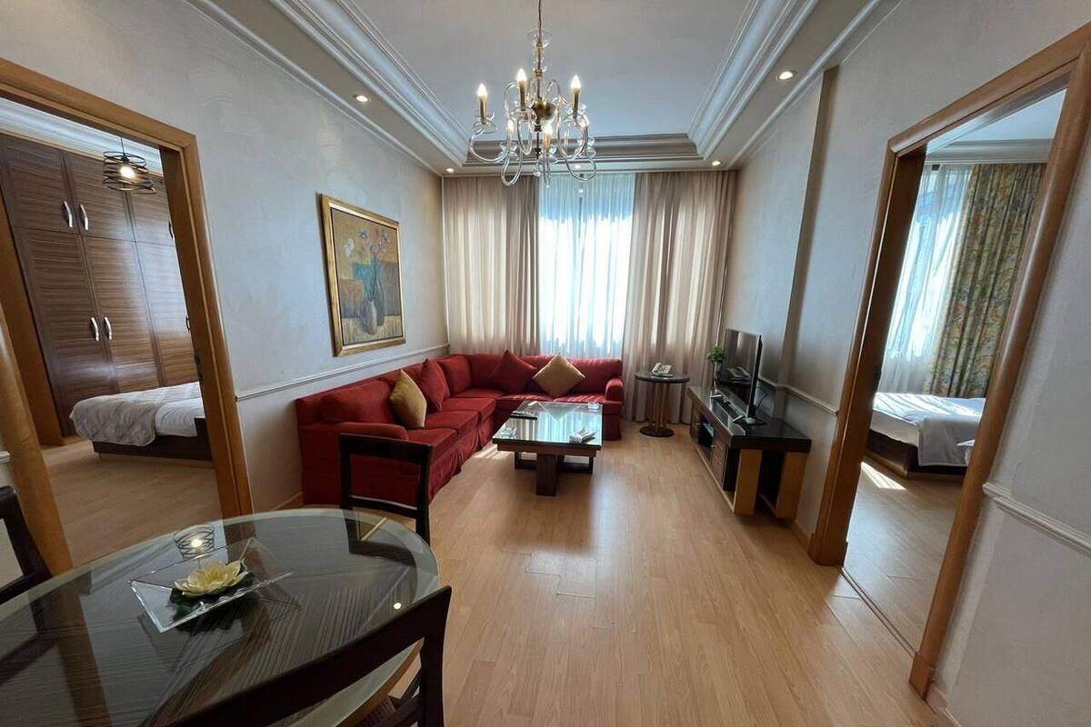 A hotel room with a wooden floor, a red sofa, a glass coffee table, a TV, and a dining area with a table and chairs.