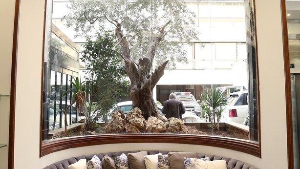 A curved grey sofa with multiple cushions, a small round table, and a large window with a view of trees and parked cars. - Verdun Suites Hotel (Beirut)