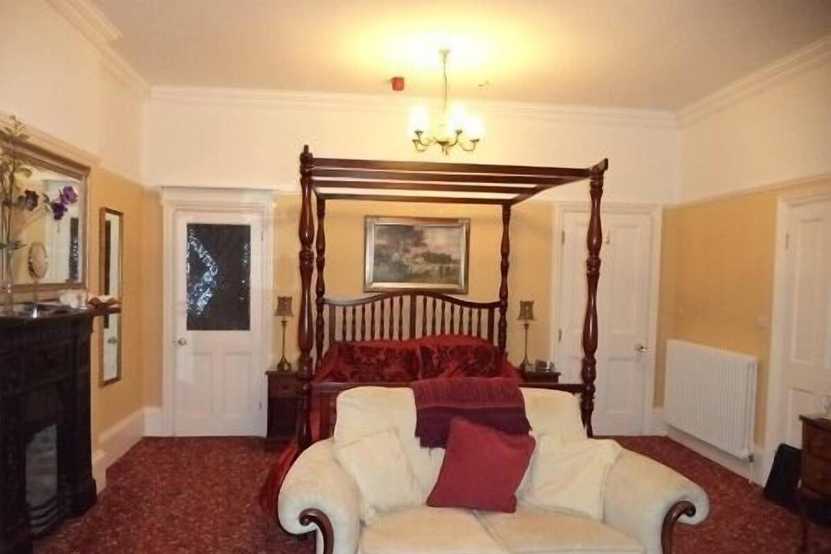 A bedroom with a four-poster bed, a white armchair, a fireplace, and a chandelier.