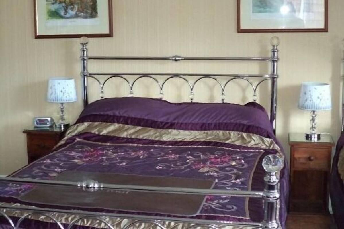 A neatly made bed with a metal frame and a purple and gold patterned bedspread. There are two bedside tables with lamps and a small clock on one.