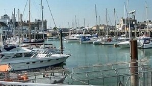 Un port de plaisance où sont amarrés de nombreux bateaux, un drapeau flottant au sommet d’un mât et des bâtiments en arrière-plan.