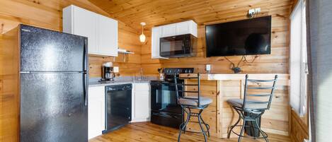 A compact kitchen with wooden walls, a black refrigerator, a microwave, a stove, and a bar stool.