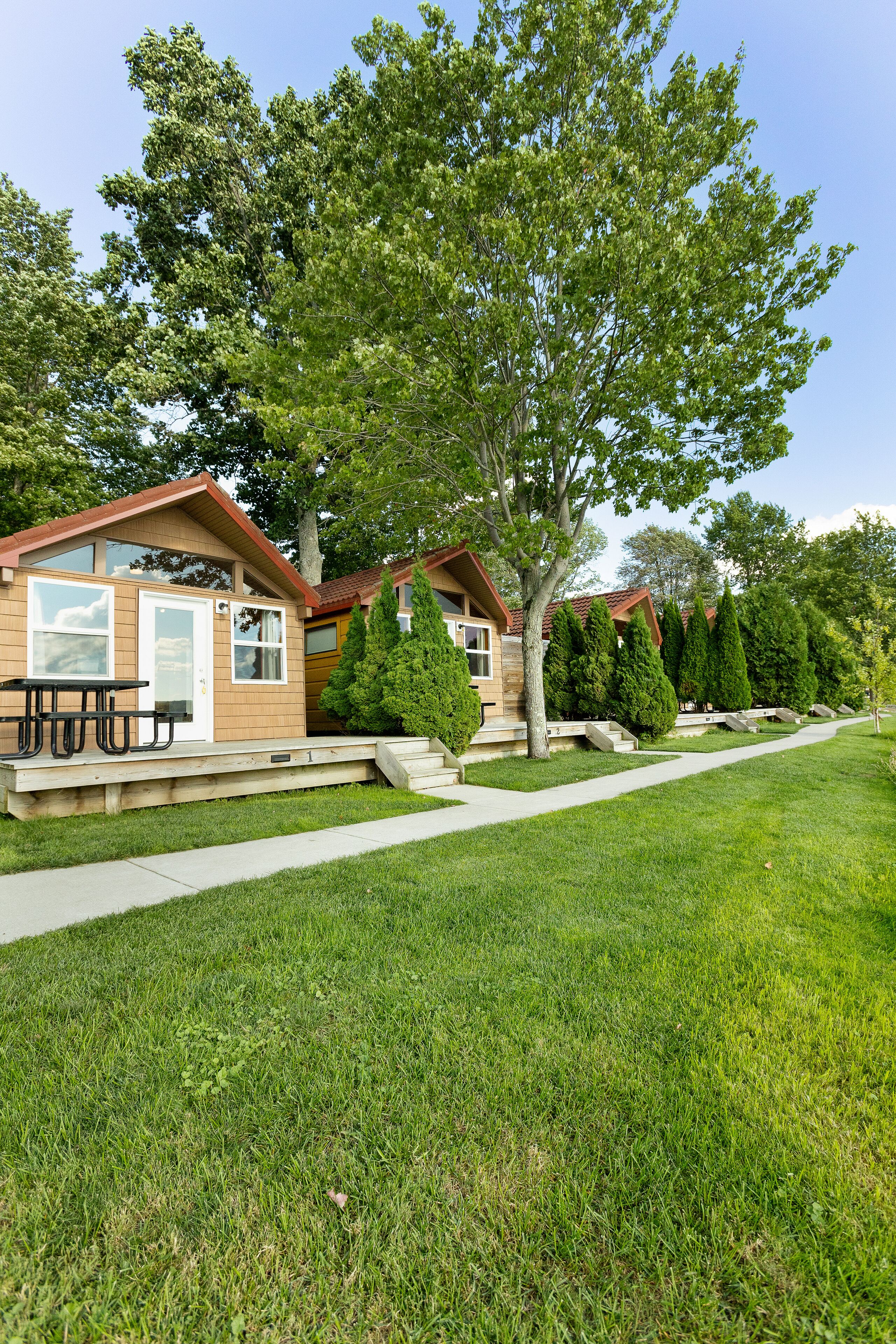 A row of wooden cabins with a picnic table in front, surrounded by well-maintained lawns and trees.