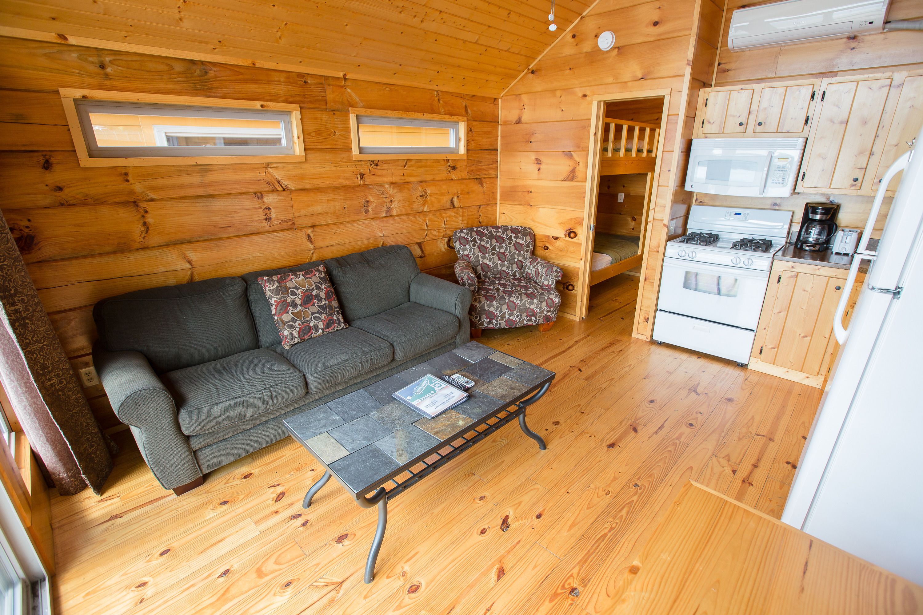 Interior acogedor de una cabaña con sofá, sillón estampado y una pequeña cocina con estufa y microondas.