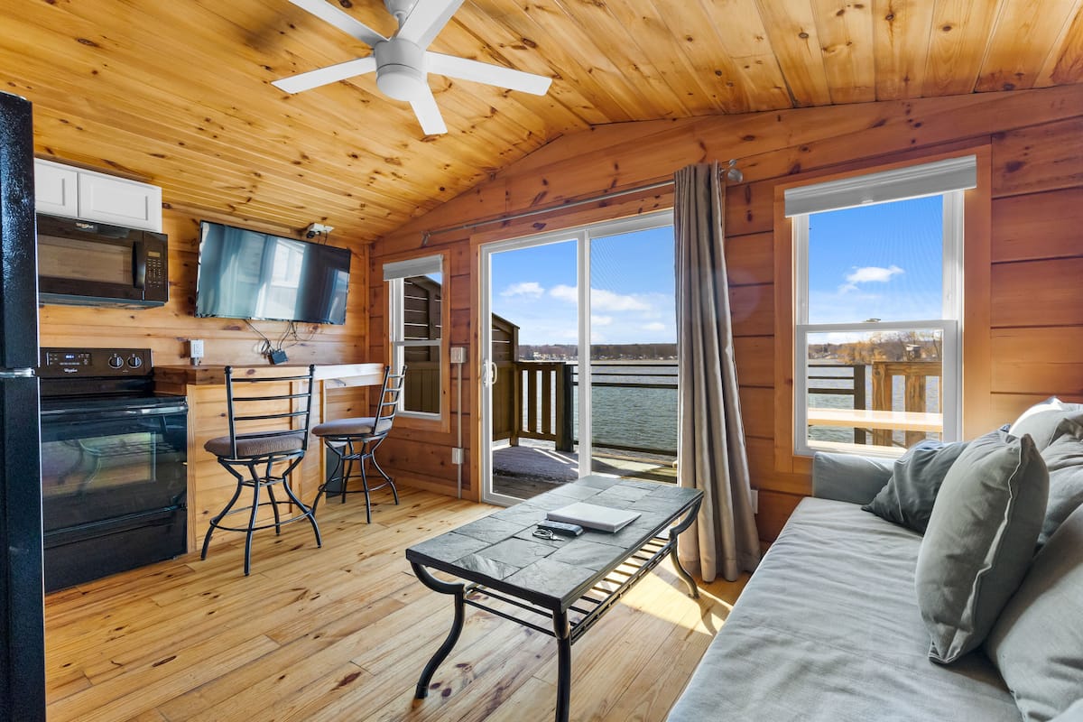 A cabin with a wooden interior, a kitchenette, a dining area, and a sofa with a view of the water.