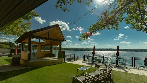 A wooden cabin with a deck overlooking a lake, furnished with outdoor seating and a table. - Edinboro Lake Resort Cabins (Edinboro)