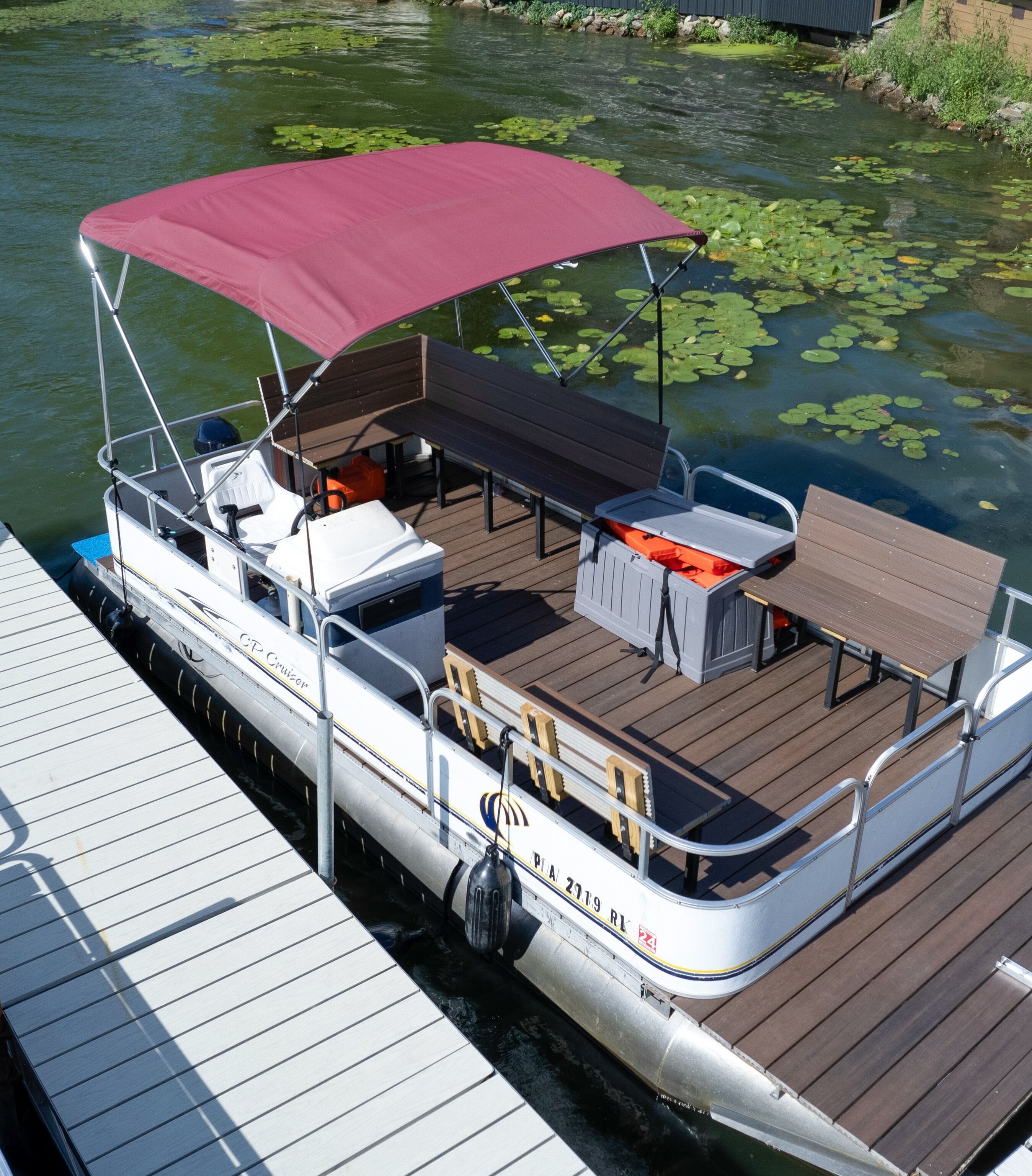 A pontoon boat docked at a pier with a covered seating area, a table, and a grill.