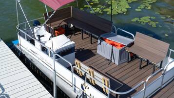 A pontoon boat docked at a pier with a covered seating area, a table, and a grill.