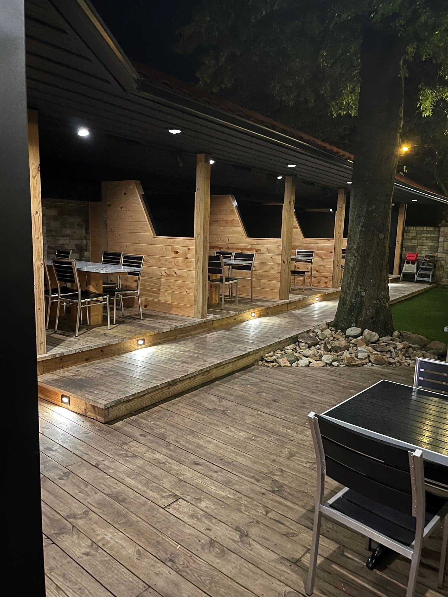 An outdoor patio area with wooden tables and chairs, a stone wall, and a tree.