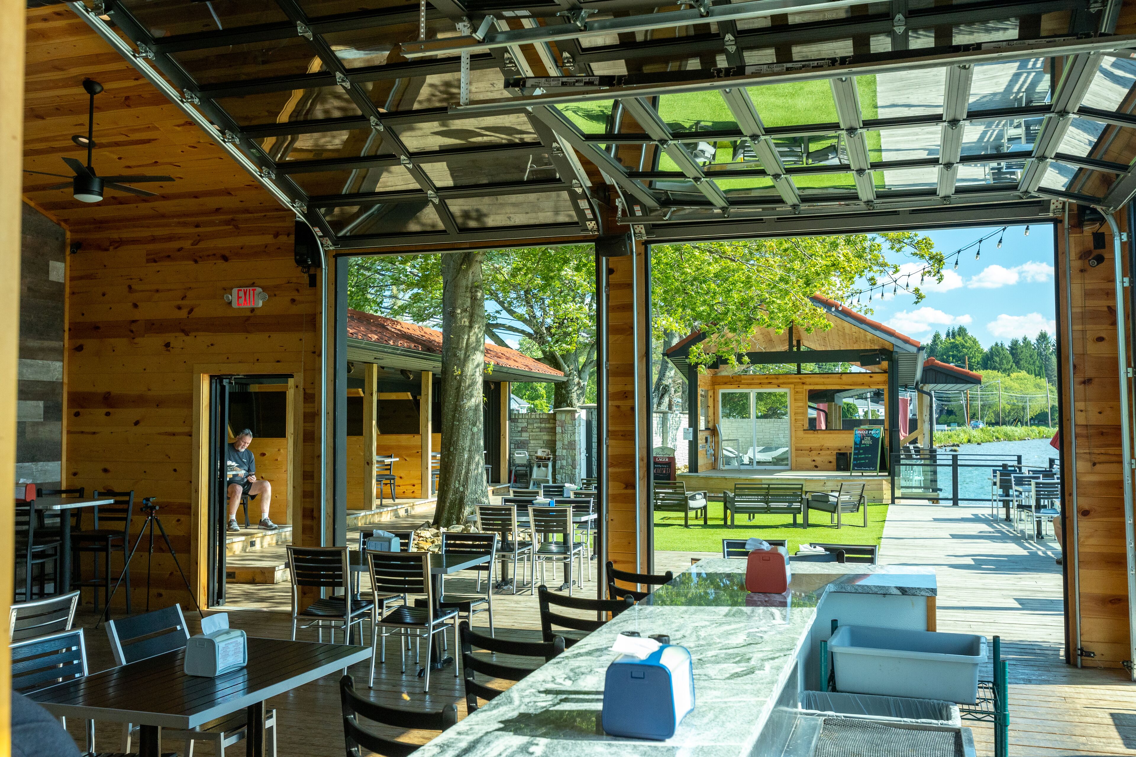 An outdoor dining area with wooden tables and chairs, a covered seating area, and a view of a building across a grassy field.