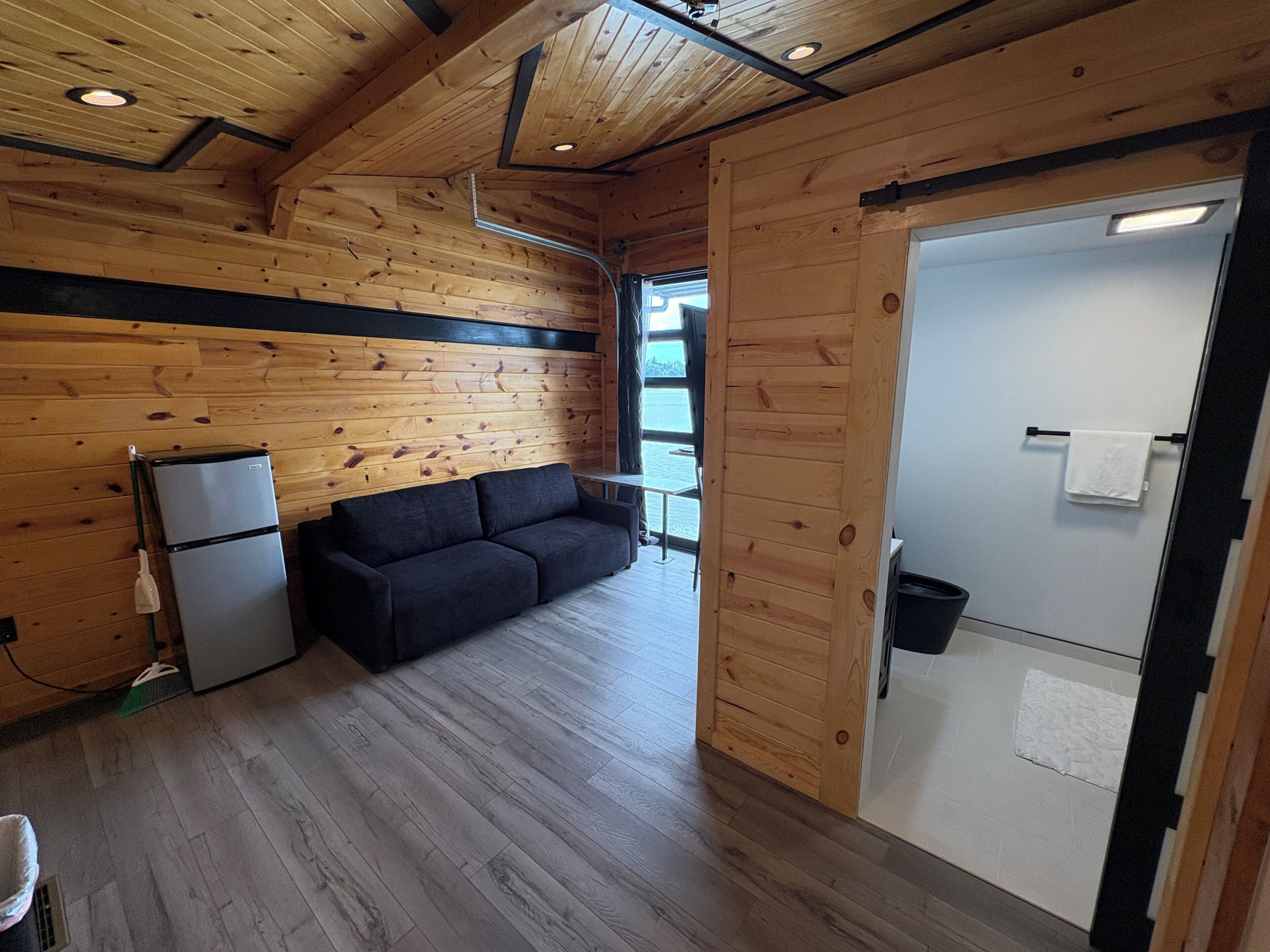 Interior de una cabaña con techo de madera, un sofá, un refrigerador y una puerta que da a un baño.