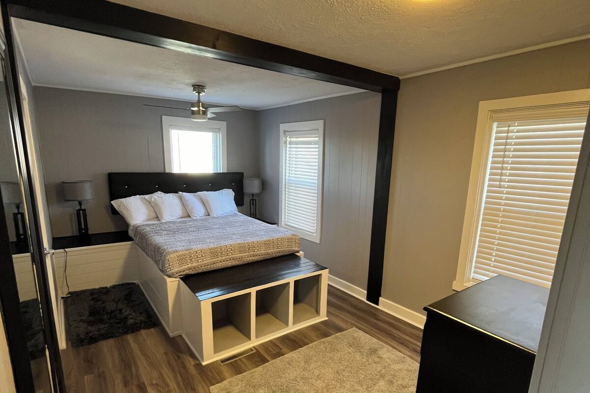 A bedroom with a bed, bedside tables, a nightstand, a ceiling light, a window with blinds, and a door.