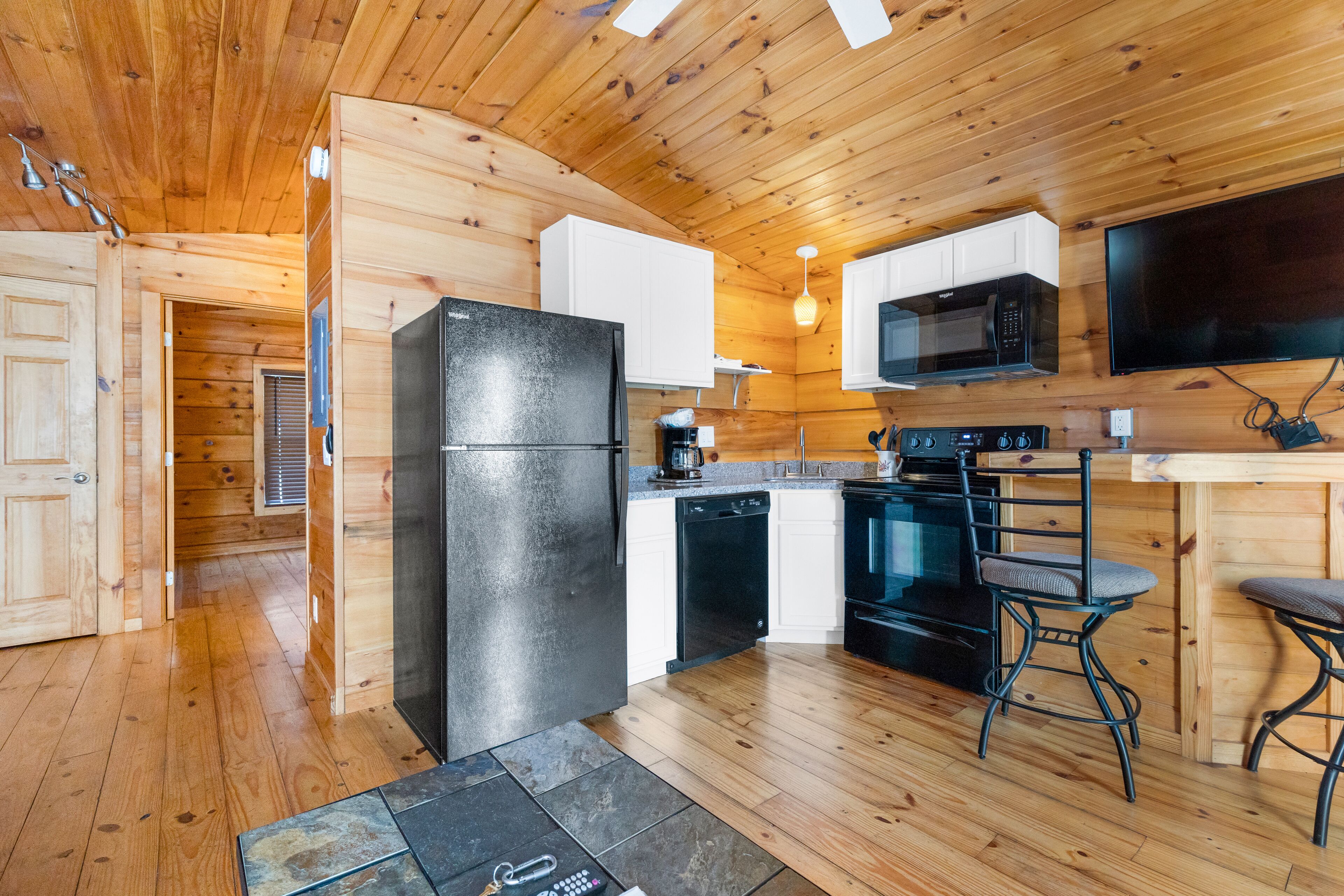 A compact kitchen with wooden walls, a black refrigerator, white cabinets, a microwave, an oven, and two bar stools.