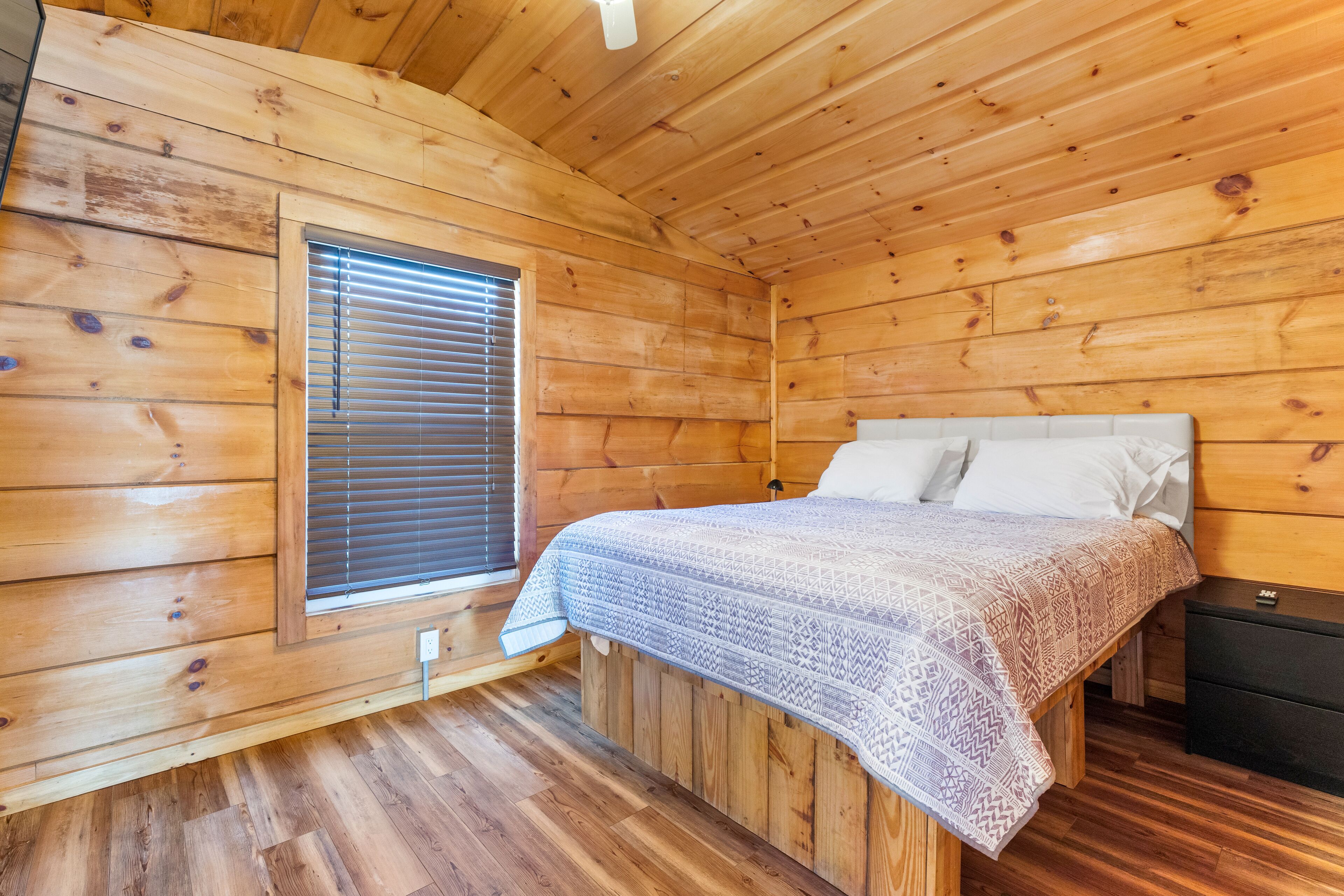 Une chambre dans une cabane en bois, avec un lit, une table de chevet et une fenêtre avec des stores.