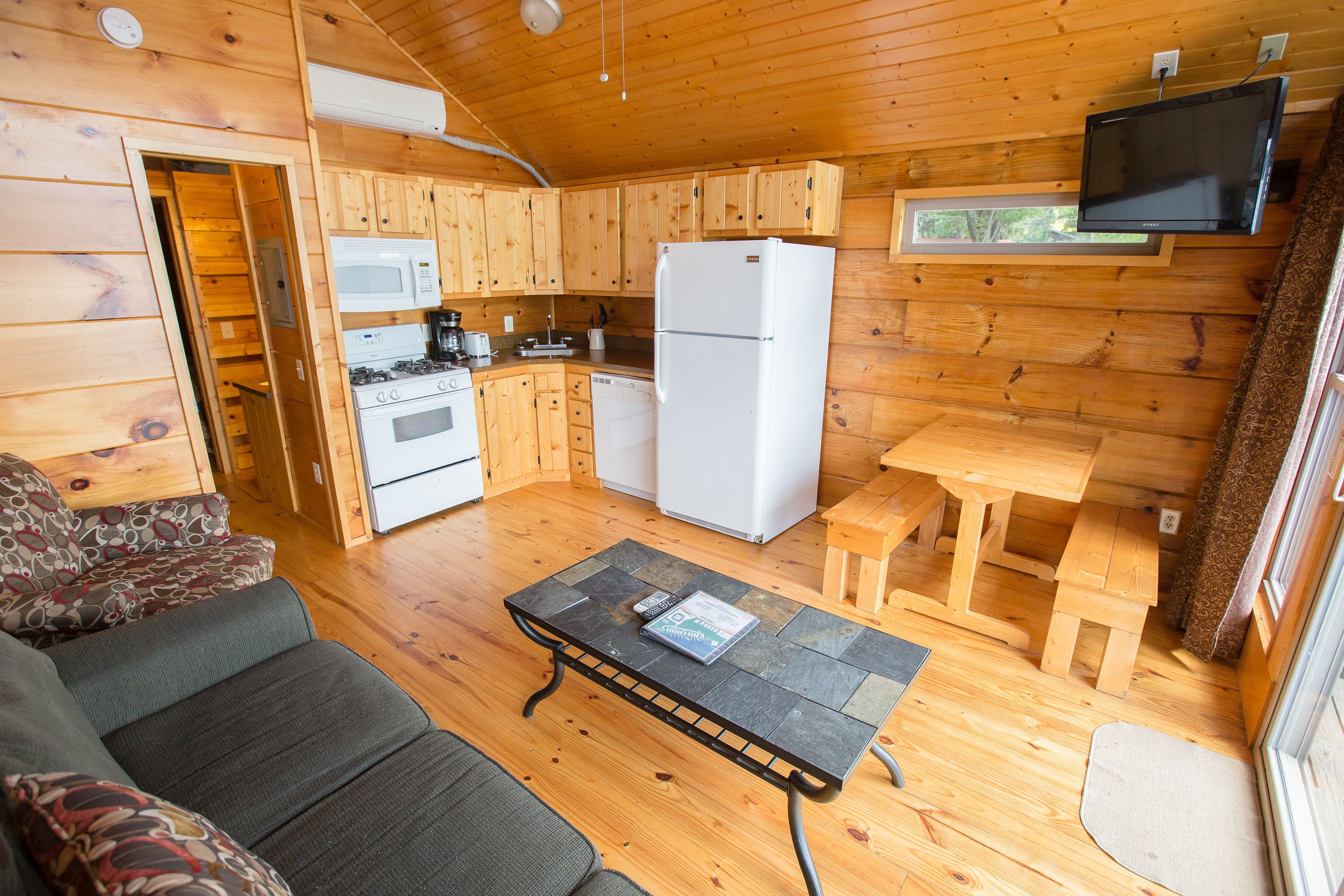 A cozy cabin-style room with a wooden interior, a sofa, a dining table, a kitchen with a refrigerator, microwave, stove, and dishwasher.
