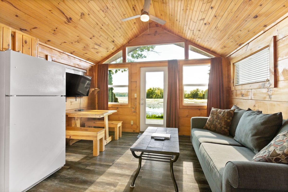 A cozy cabin interior with a wooden ceiling, a kitchenette, a dining area, and a sofa with cushions.