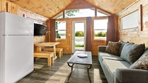 A cozy cabin interior with a wooden ceiling, a kitchenette, a dining area, and a sofa with cushions. - Edinboro Lake Resort Cabins (Edinboro)