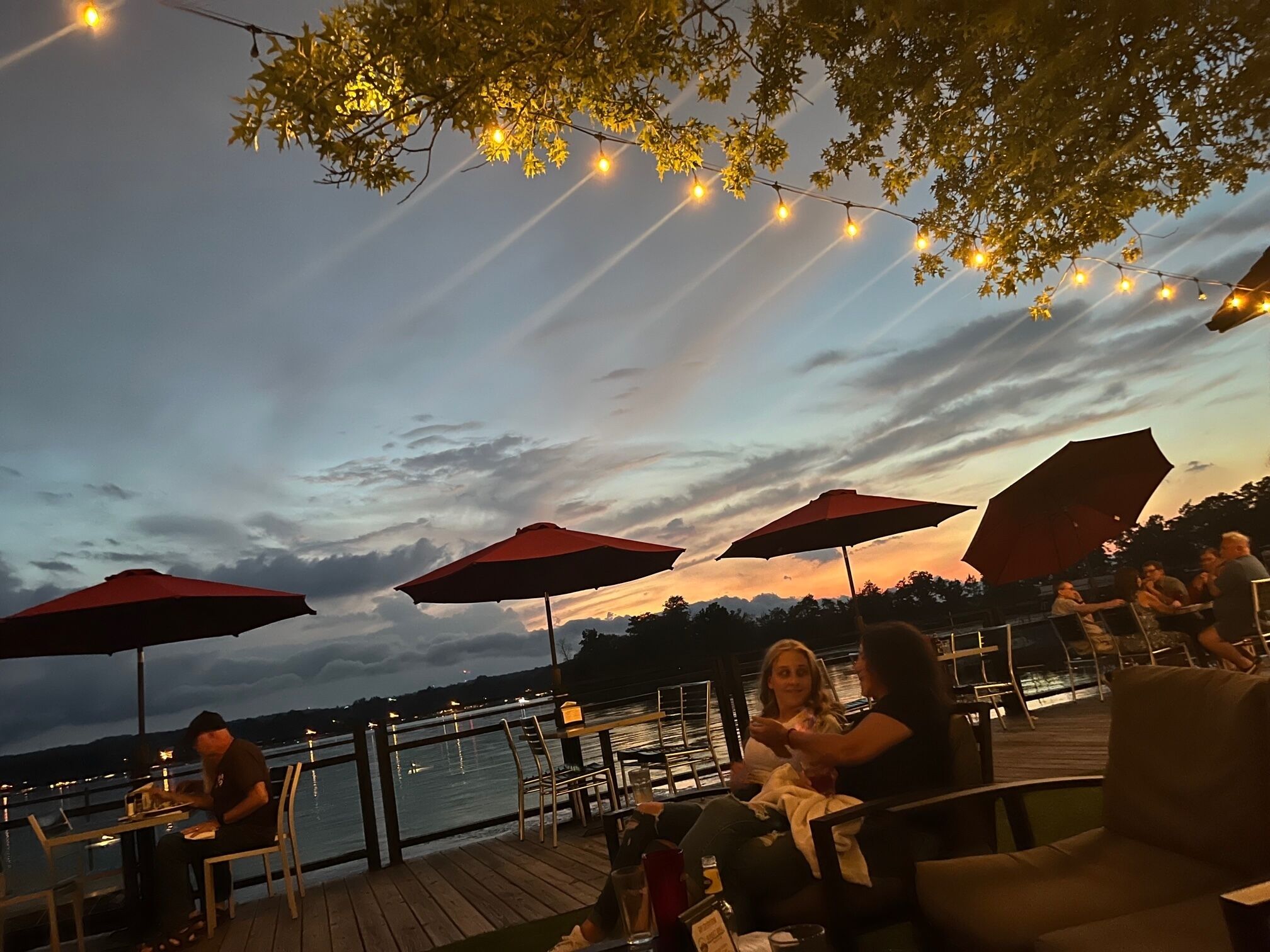 Un espacio de comedor al aire libre con mesas y sillas, sombrillas y luces colgantes, con el atardecer sobre el agua de fondo.