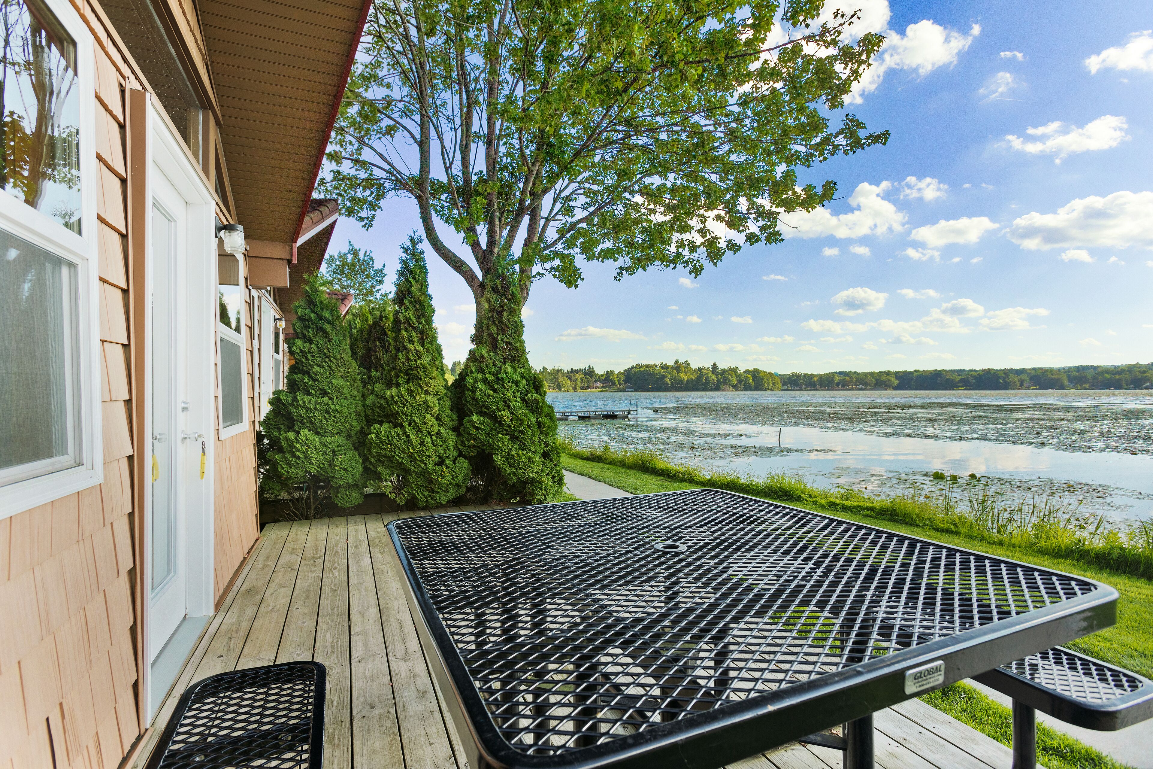 A wooden deck with a metal table and bench, overlooking a lake with trees and a clear sky.