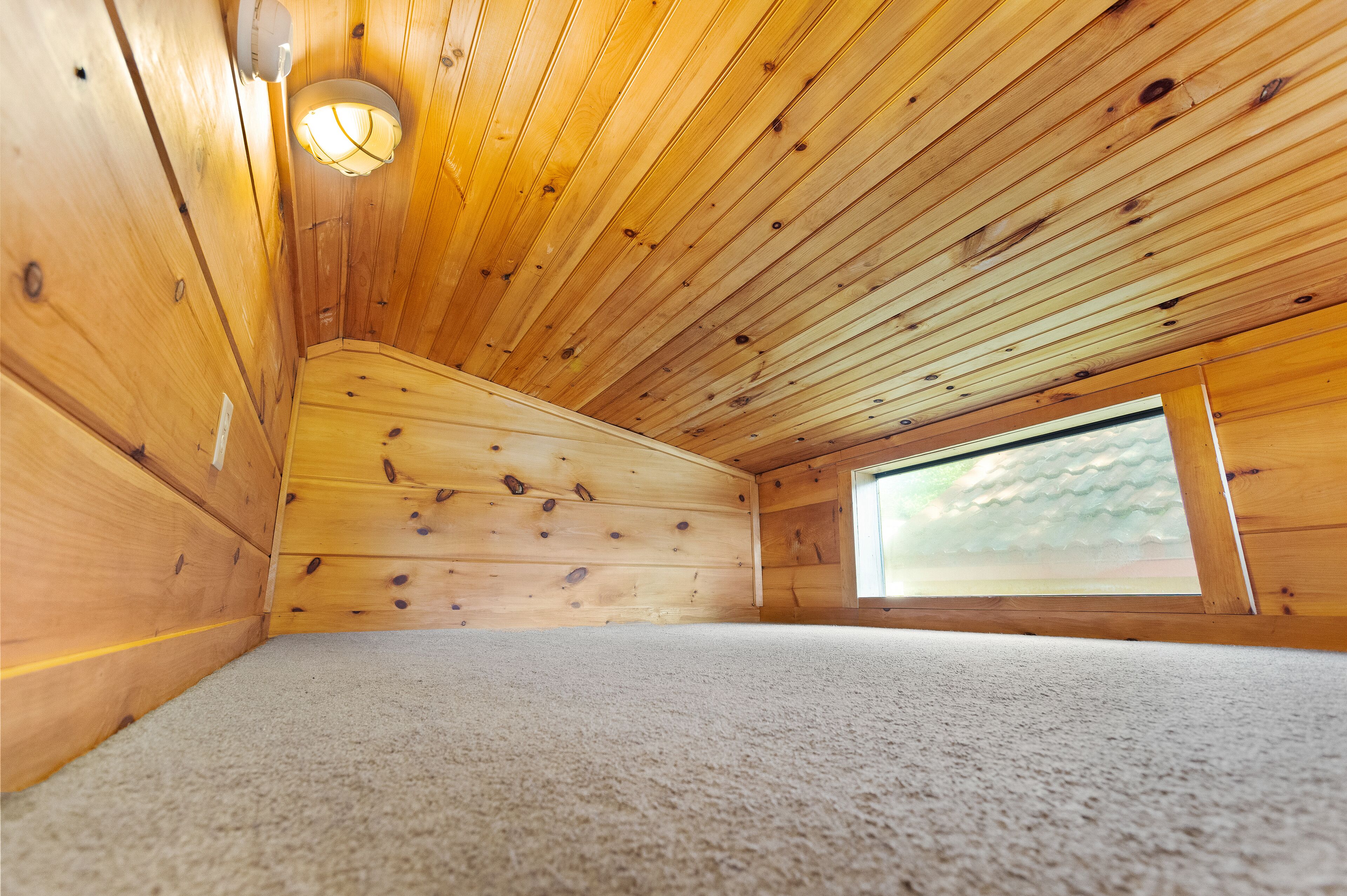 A small, wooden attic room with a sloped ceiling, a single window, and a carpeted floor.