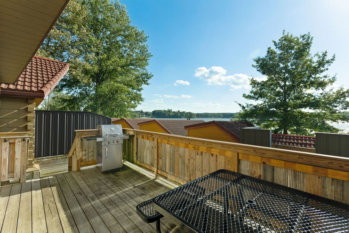 A wooden deck with a grill and a table, overlooking a body of water and trees.