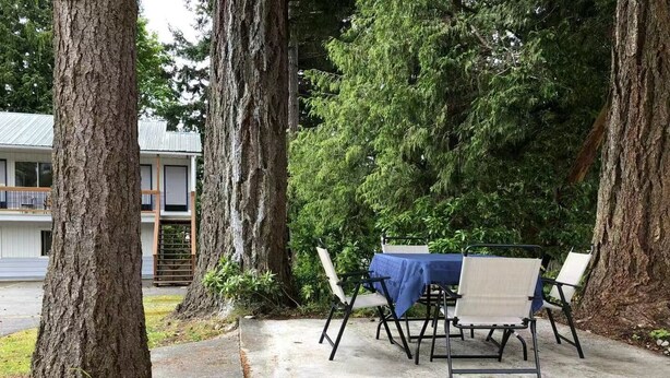 An outdoor patio with a table and chairs, surrounded by trees and a building in the background. - Marland Motel (Powell River)