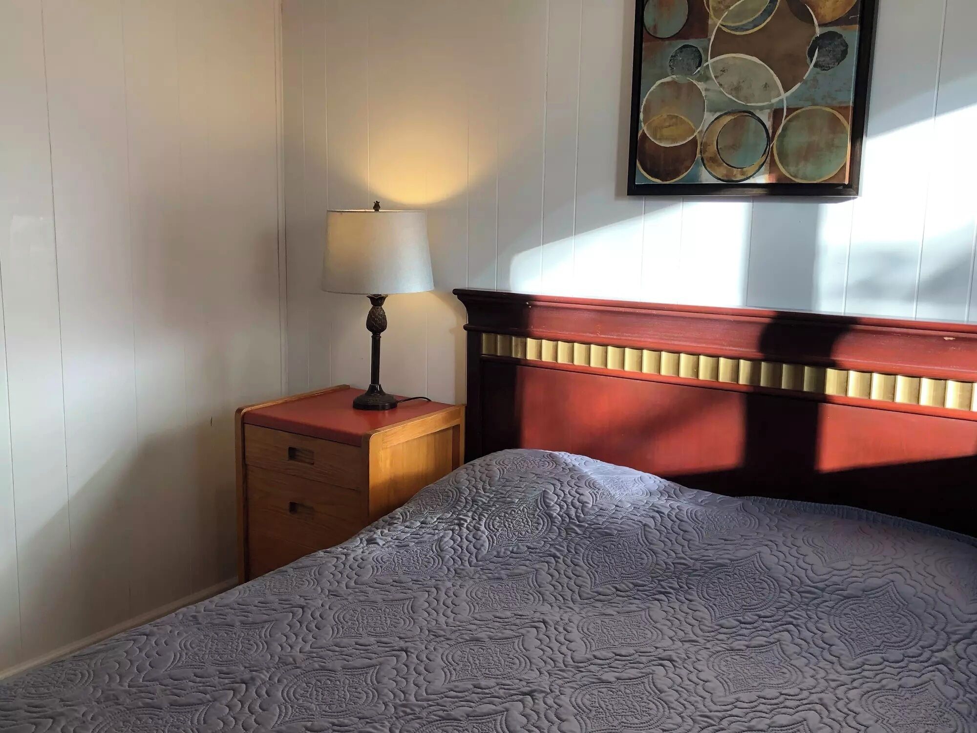 Een keurig opgemaakt bed met een beddekleed met dessin, een nachtkastje met een lamp, en een aan de muur bevestigd kunstwerk.
