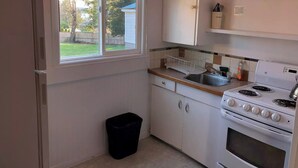A compact kitchen with white cabinets, a stove, and a window overlooking a backyard. - Marland Motel (Powell River)