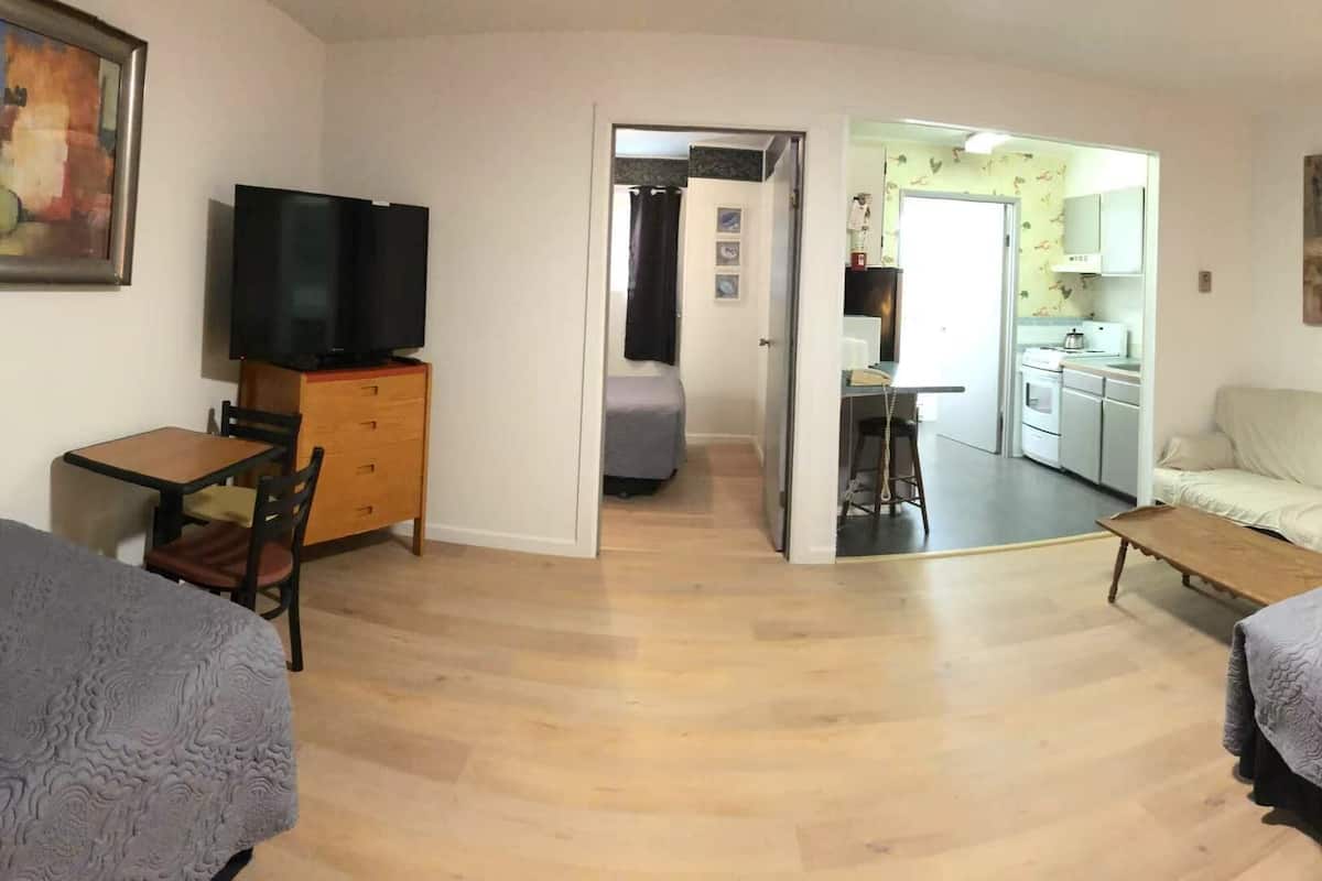A compact hotel room with two beds, a television on a wooden cabinet, a small table, and a kitchenette in the background.